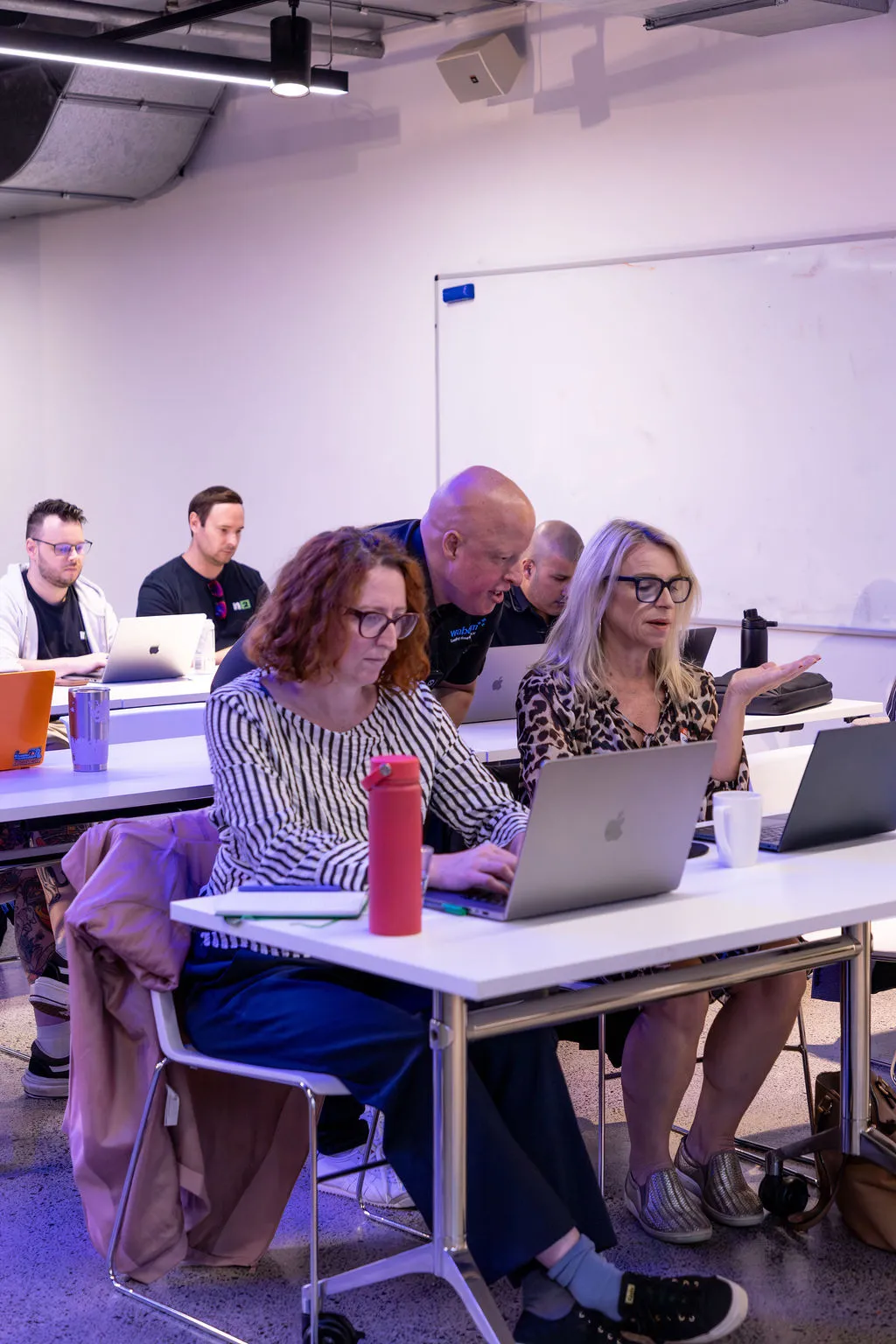 Two women building their own AI tools on their laptops, with Leo helping at their table